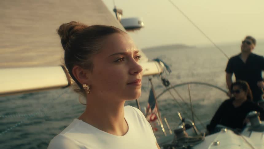 Two young women ride on a sailboat at sunset the ocean glowing warm orange around them while a young man in sunglasses stands at the wheel turning it as the boat cuts smoothly through the water