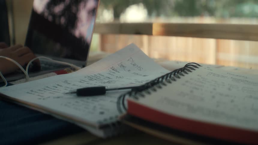 A page blows in the wind and shows handwritten homework and lecture notes in a notebook placed beside it in the bright sunlight