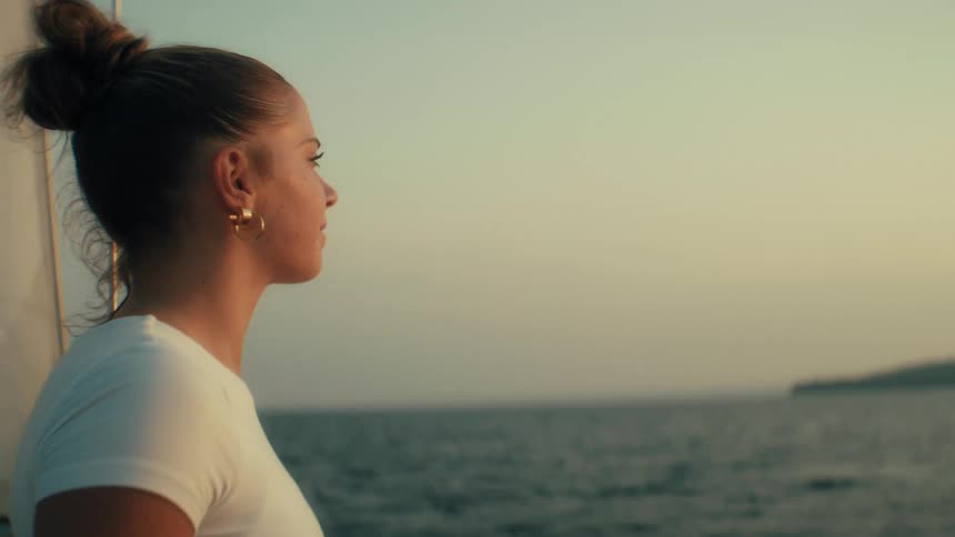 Woman in white shirt and gold earrings gazing at the sea during sunset on sailboat in contemplative moment