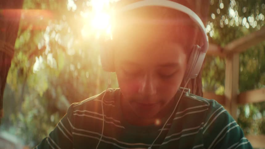A teenage boy sits in a halfbuilt tree house in the woods at sunset putting on a wired headphones as warm orange light filters through the trees