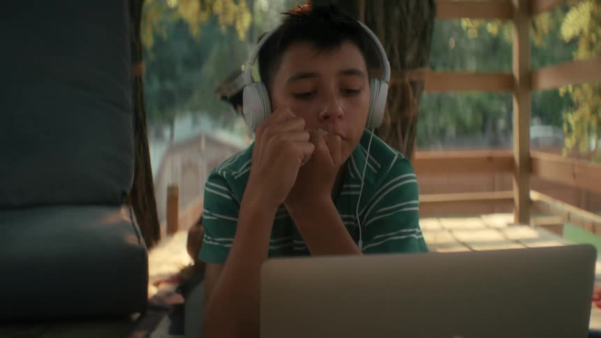 Boy on a deck writes in a notebook then looks at his laptop while wearing headphones and listening to a classroom lesson and lecture