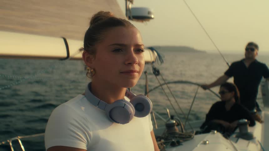 Teenager girl wearing a white blouse removing her headphones and looking around while sitting on a sailboat on the ocean next to two other people on the background outdoors