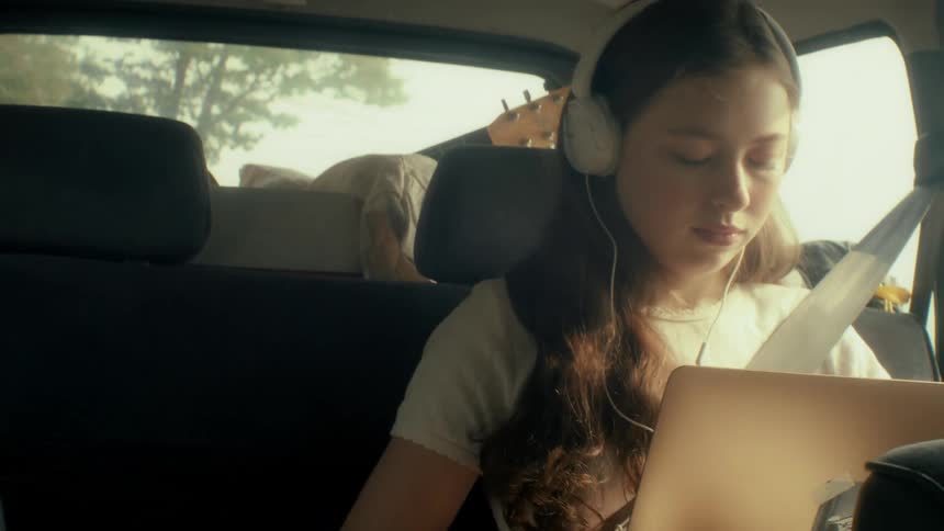 A teenage girl sits in the back seat of a moving car wearing headphones and looking down at a laptop while other passenger is looking at the window