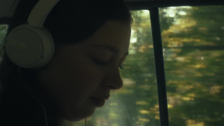 A young woman with her hair in a bun wears white overear headphones and gazes out a car window at passing trees looking forward with a calm and reflective mood