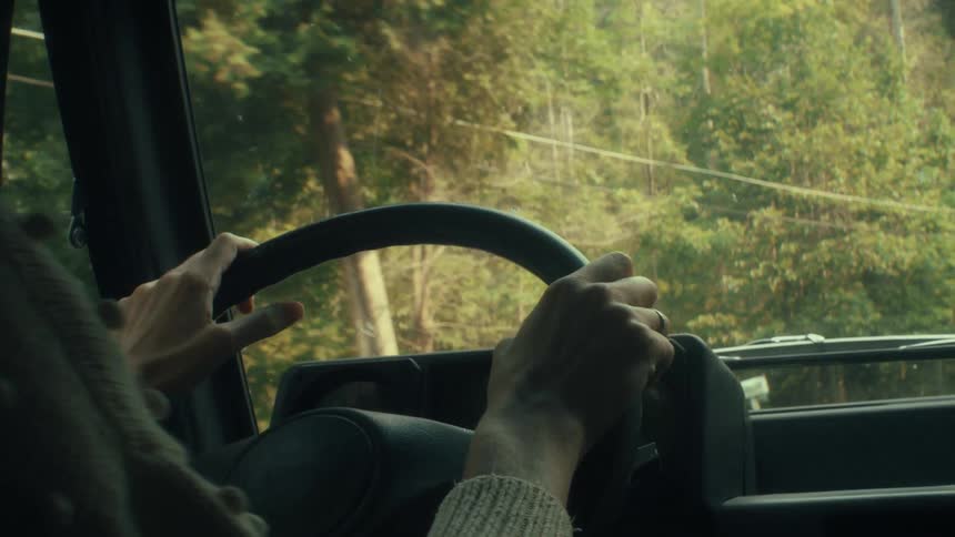A woman wearing a beige sweater and a ring drives a car along a road lined with trees