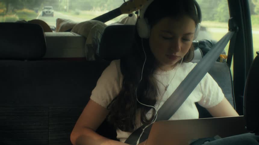 Teenage girl typing on the keyboard of a laptop computer as she sits in the back seat of an automobile