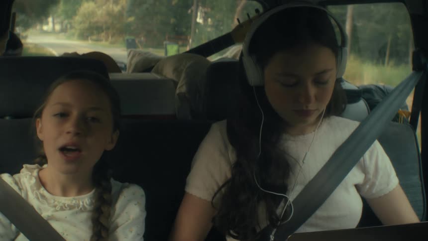 Two girls sit in the back of a moving car during a road trip the younger girl sings while the older girl wears headphones and looks down at a laptop as sunlight filters through the windows and trees pass by outside
