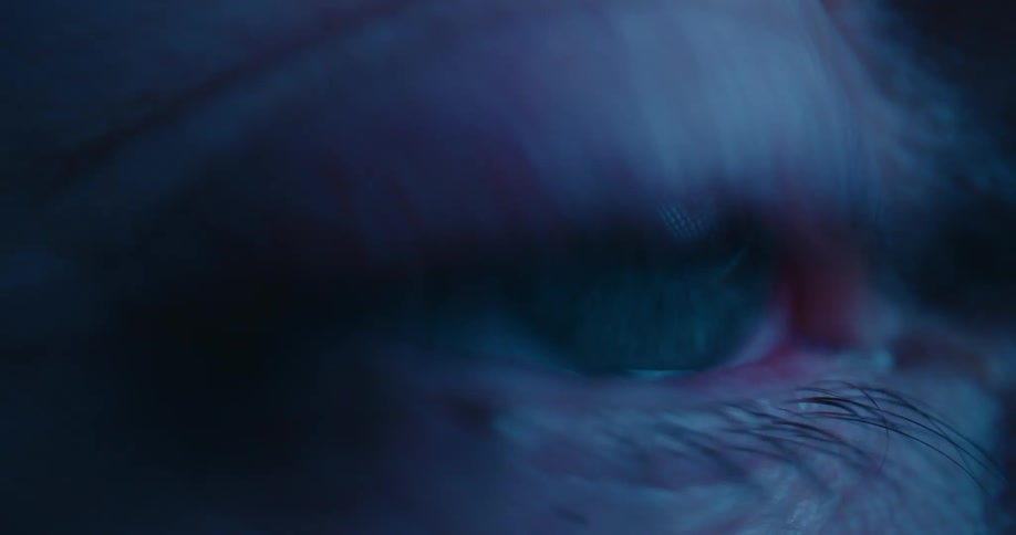 A human eye focuses on a glowing screen the shifting light casting soft reflections across the face the eyelid blinks several times
