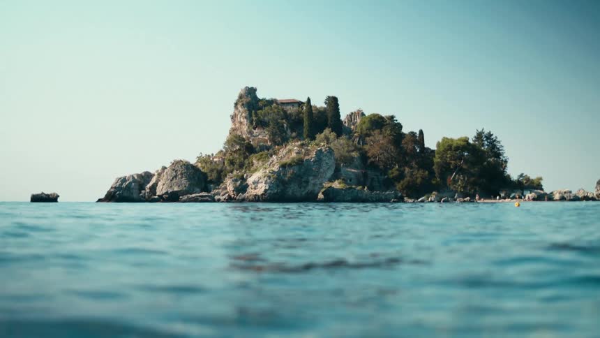 A small rocky island sits alone in the middle of the ocean at midday bright sunlight hitting the rough stone as waves move around it