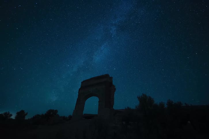 A breathtaking timelapse captures the milky way and countless stars rotating across the night sky above a silhouetted ancient ruin before fading into the bright blue of dawn