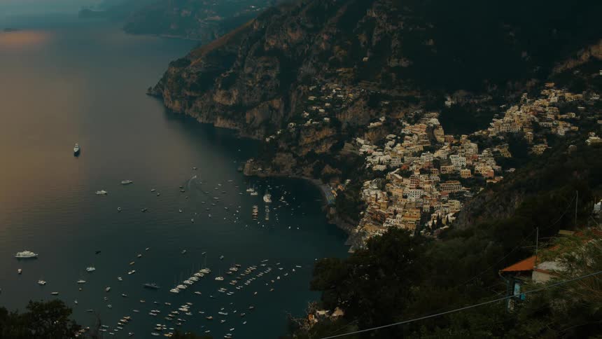 A coastal town sits high on the hillside at sunset overlooking an ocean bay where sailboats are docked their masts catching the warm orange light as the sky fades into evening