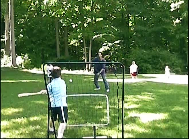 Boys playing baseball with a young adult man in a yard surrounded by tall trees with a house on the background outdoors during day time