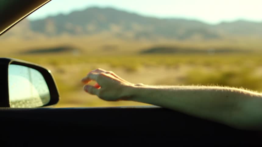 Arm extended out of a moving car window with blurred rural landscape in the background