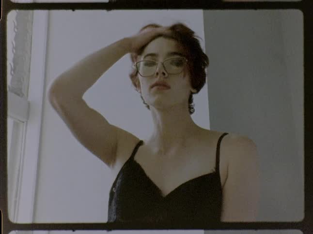 Person wearing glasses and a black top rests a hand on the face beside a window indoors