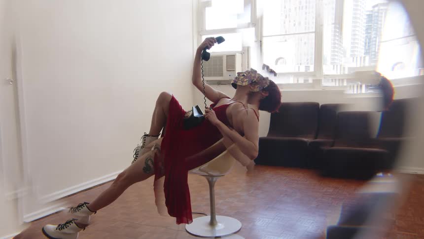 Woman reclining on a swivel chair holds a corded telephone wearing a red dress in an indoor space with a window in the background