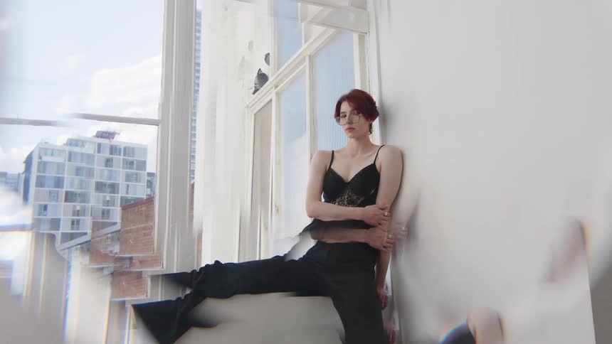 Woman leaning against a wall sits on a window ledge with legs extended and buildings visible in the background