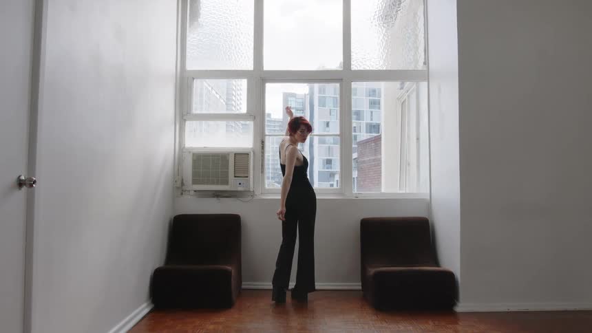 Woman standing in front of a window touches her hair wearing black clothing in an indoor space with buildings in the background