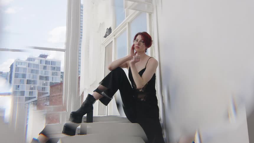 Woman sitting on a window ledge raises one arm and touches her face wearing black clothing in an indoor space with buildings in the background