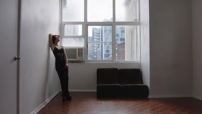 Woman leaning against a wall tilts her head back in front of a window in an indoor space with buildings in the background
