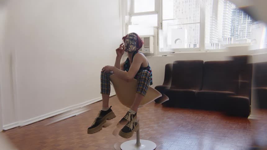 Woman seated on a swivel chair rests her arms on her legs wearing patterned clothing and a mask in an indoor