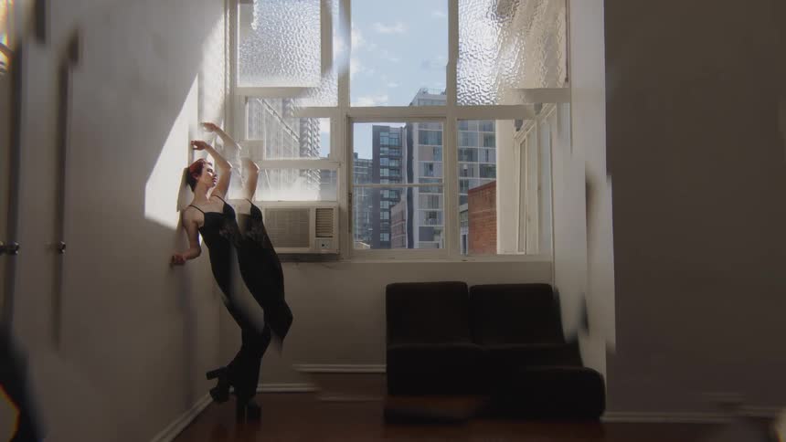 Woman leans her body backward with one leg raised in front of a window in an indoor space with buildings in the background