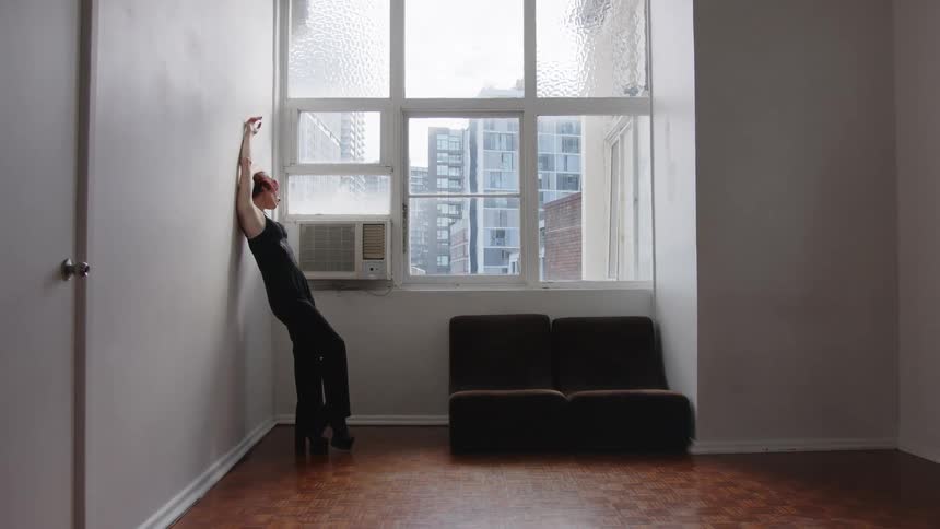 Woman leaning against a wall tilts her body beside a window in an indoor space with buildings in the background