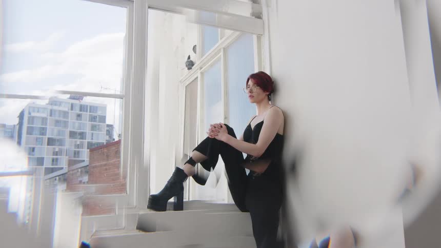 Woman sitting on a window ledge hugs her legs wearing black clothing in an indoor space with buildings in the background