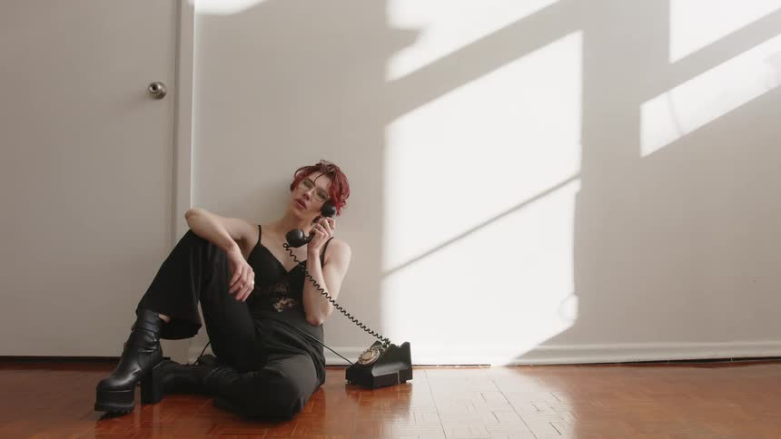 Woman sitting on the floor holds a corded telephone beside her body in an indoor space with natural light