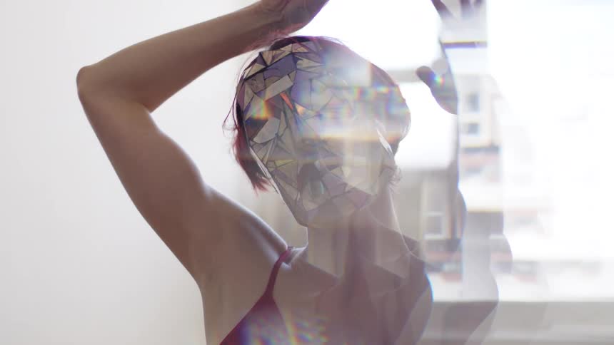 A woman with short red hair wearing a red top and a reflective mosaic mask moves her arms and head while a kaleidoscopic lens effect creates shimmering light patterns