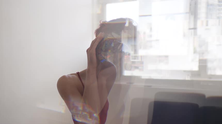 Woman wears a geometric mask and partially covers her face with her hand in an indoor space with a window in the background
