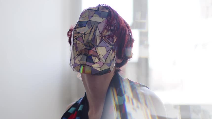A young woman with red hair sits in a room as bright daylight shines through the window she wears a mask and slowly moves her face and shoulders