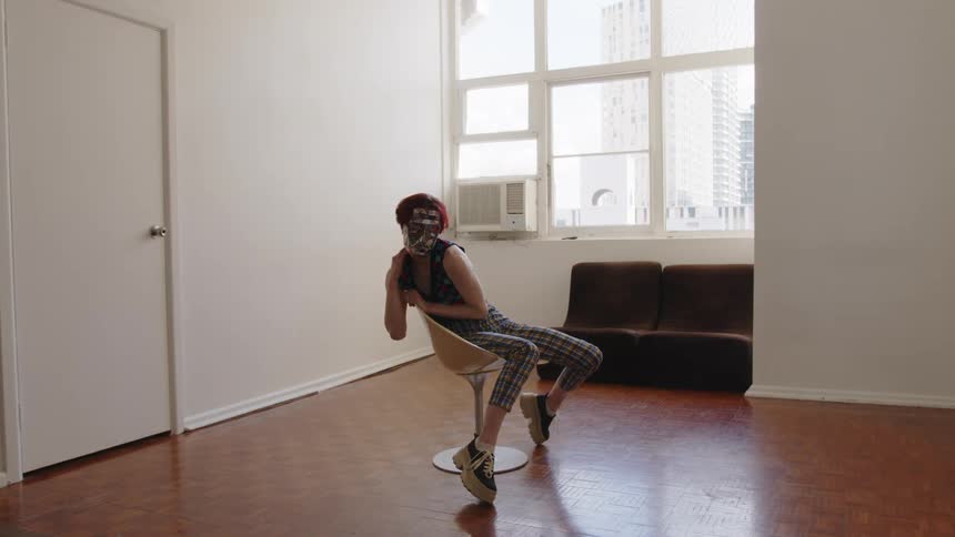 Woman seated on a swivel chair tilts her head wearing patterned clothing and a mask in an indoor space with a window in the background