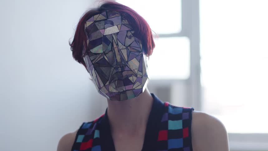 Woman wears a colorful geometric mask and patterned clothing in an indoor space with a window in the background