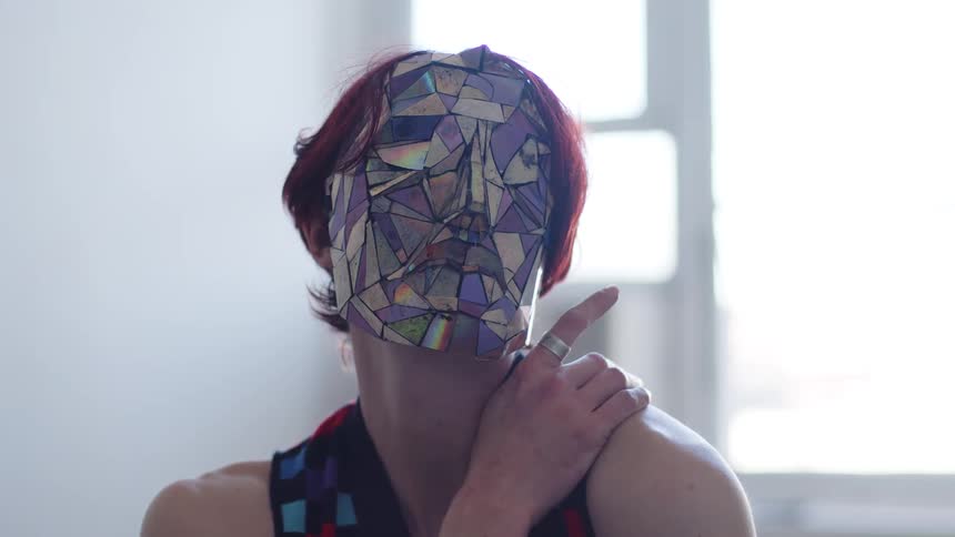 A young woman with red hair sits in a welllit room at midday wearing a mask and a dress sunlight from the window brightens the space as she slowly moves her head