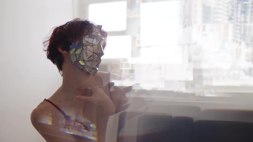 In a medium shot with a double exposure effect a person with short red hair wears a mask made of reflective geometric shards and a thin strapped top while moving their arms and hands gracefully against a bright blurred city background
