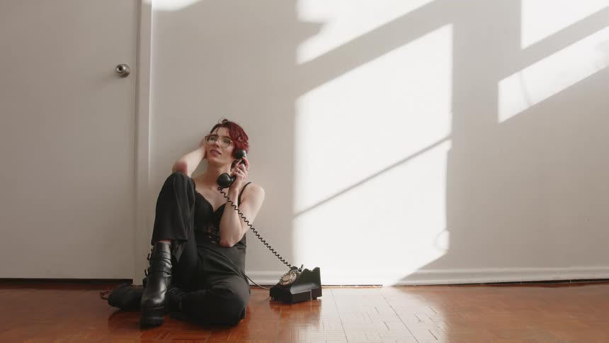 A woman sits on a wooden floor in a patch of bright sunlight leaning against a wall while listening intently to a conversation on a vintage rotary telephone