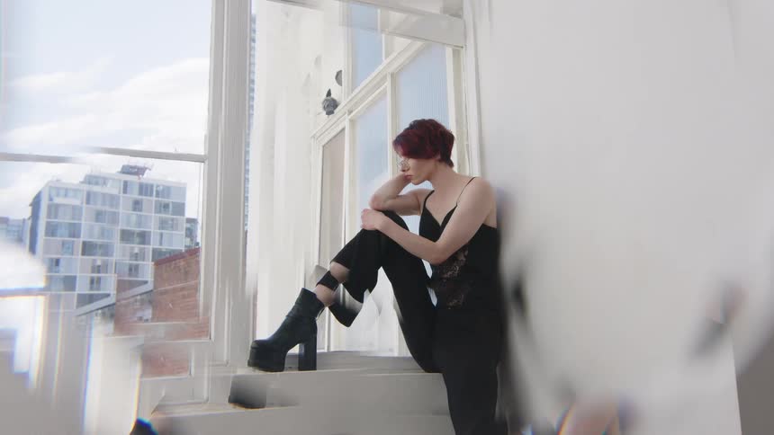 Woman sitting on a window ledge rests her head on one hand wearing black clothing in an indoor space with buildings in the background