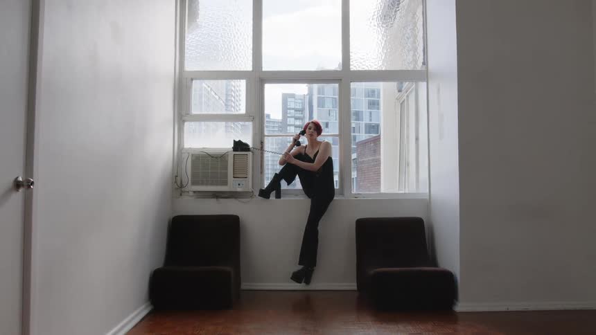 A woman with short red hair wearing a dark jumpsuit and platform boots sits on a windowsill and talks on a vintage telephone in a room overlooking city buildings