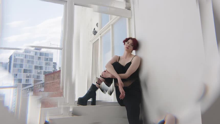 Woman sitting on a window ledge crosses one leg and rests her arms in an indoor space with buildings in the background