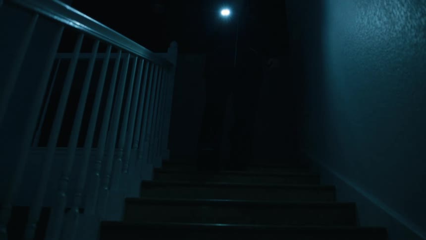 A man holding a bright flashlight walks down a dark wooden staircase at night his body mostly obscured by shadows as he descends toward the camera while a white railing and textured wall are visible in the dim bluetoned light