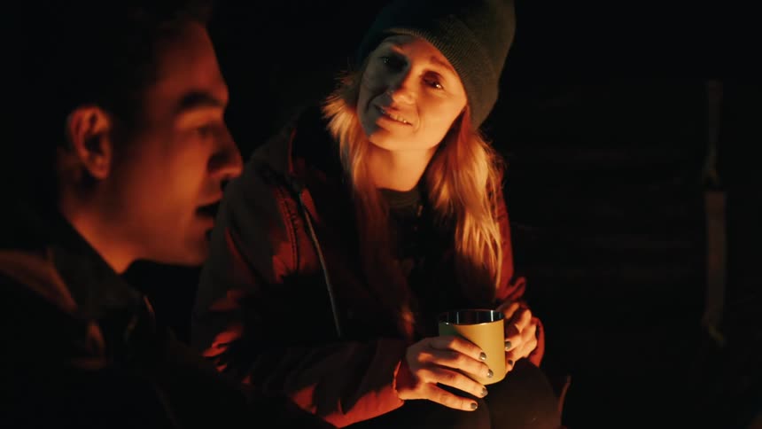 Woman in a dark environment holds a metal mug while looking at a man under warm lighting