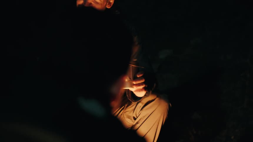 Young adult man wearing a cap sitting down in front of a camp fire talking and laughing in front of another man outdoors during night time