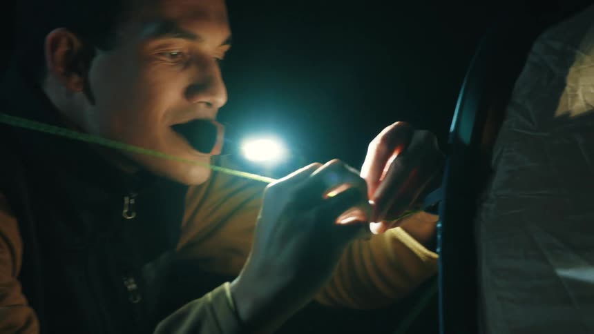 A man holds a light in his mouth as he ties a tent string in the dark