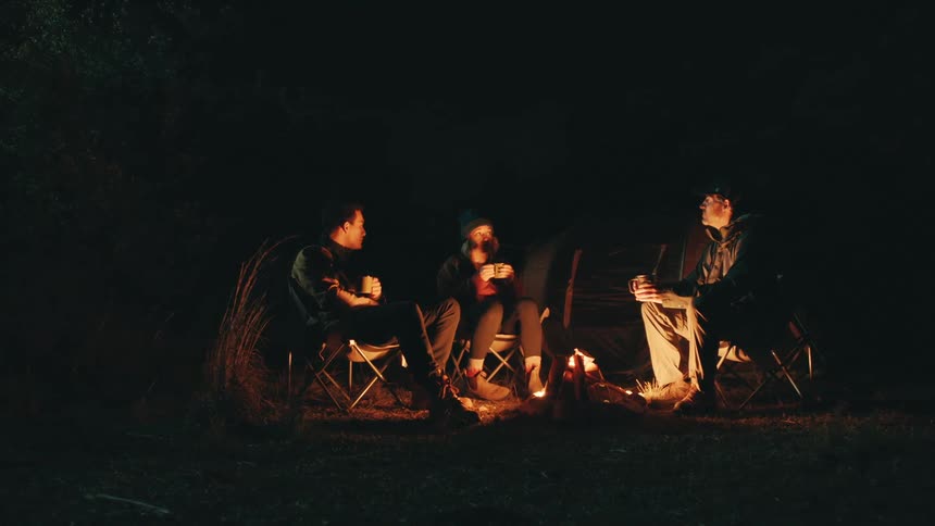 Three campers sitting in chairs illuminated with candle light holding mugs and talking