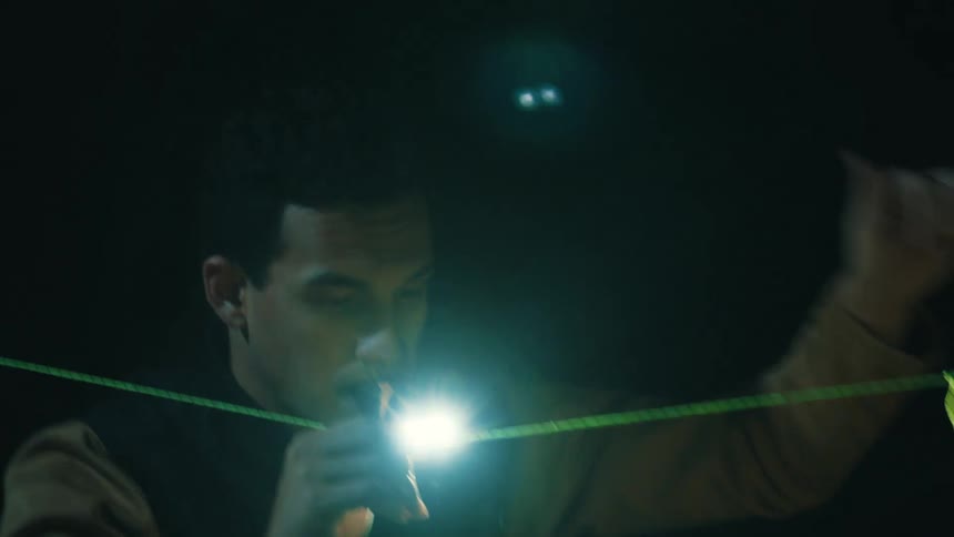 Young adult man wearing a yellow jacket pitching a tent while holding a flashlight on his mouth and making a thumbs up sign with his hand outdoors during night time