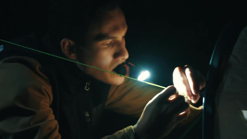Illuminated by a small flashlight held in his mouth a man carefully threads a thin rope with his hands concentrating on setting up his tent in the pitchblack darkness