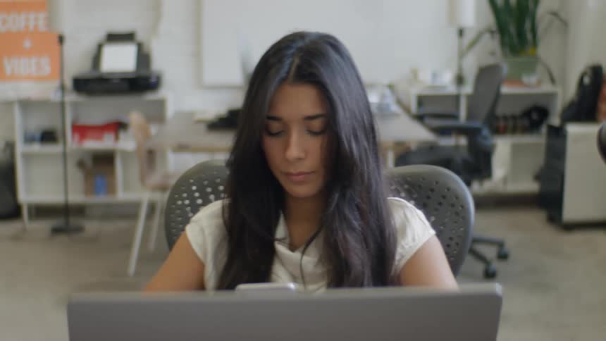 A woman with long dark hair wearing a white shirt sits at a desk in an office and looks down at a mobile phone