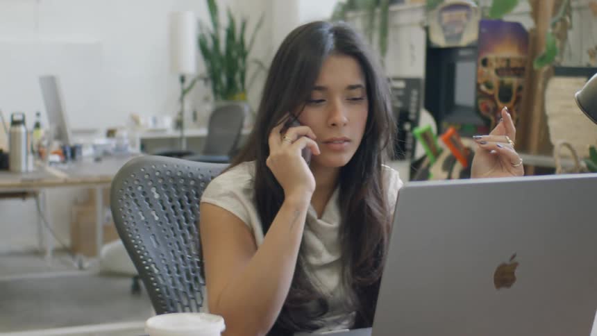 A woman with long dark hair wearing a white shirt speaks on a mobile phone while working at a laptop in a bright office