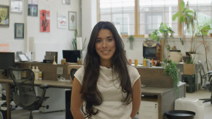 Woman in an office environment stands in front of work desks computers and plants