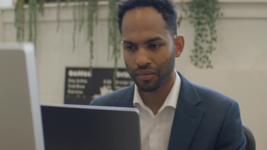 A man in a navy blazer and white shirt sits at a desk focused intently on his laptop screenhe maintains a serious expression eventually resting his hand on his chin in a thoughtful or pensive gesture